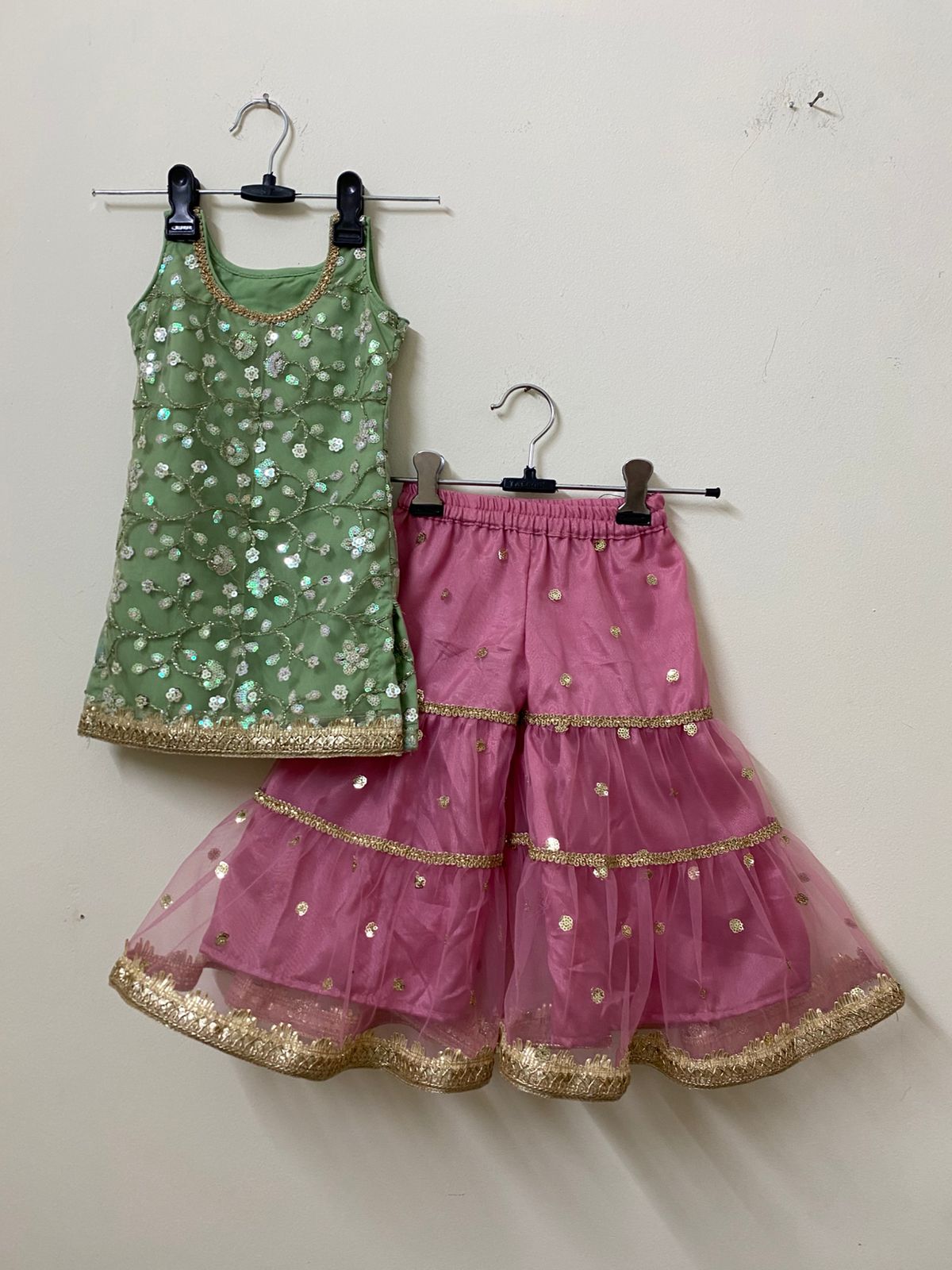 Green top and pink skirt with gold details on hangers against a white wall.